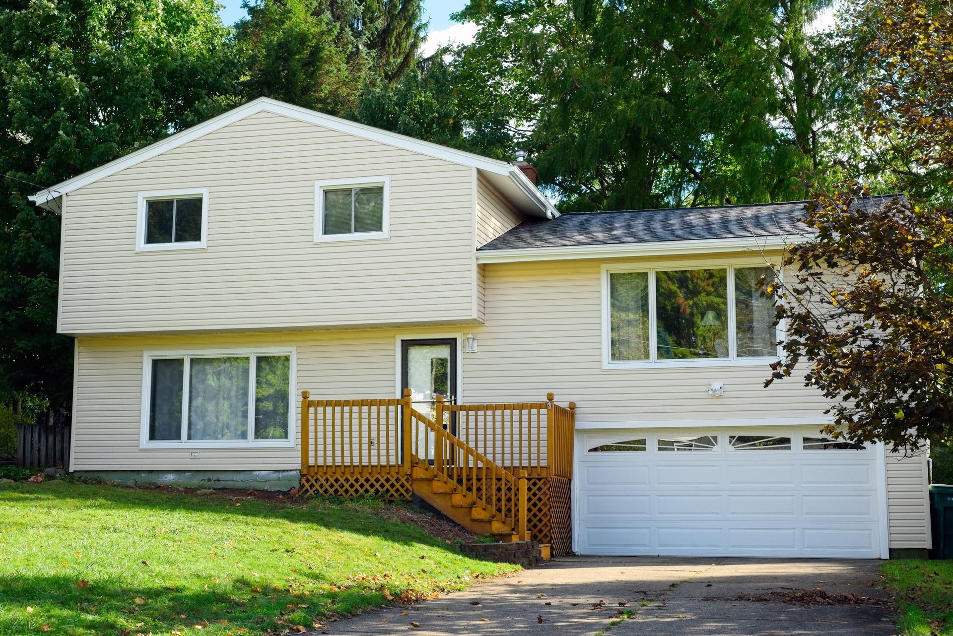 Beige two-story house with a wooden deck, white garage door, and green lawn.