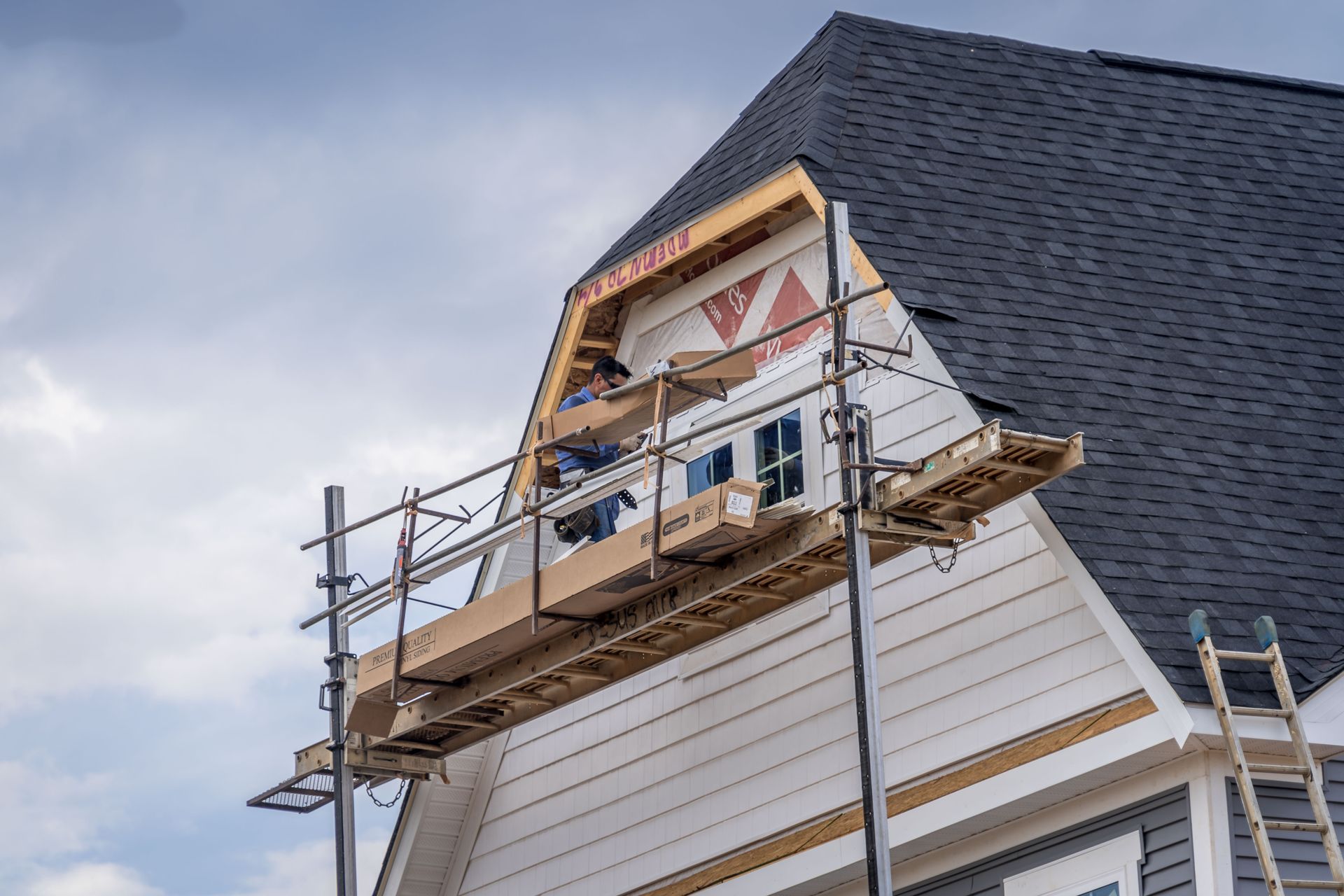Construction workers on a scaffold installing siding on a house under a cloudy sky.