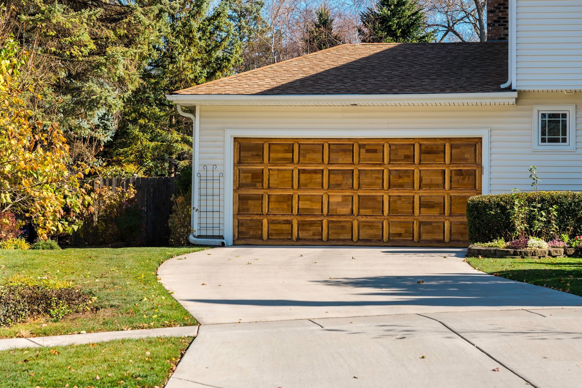 Garage with wooden door, driveway, and lawn. House with white siding, and trees in background.