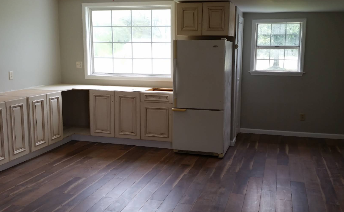 A kitchen with wooden floor, windows, white cabinets and a white fridge.