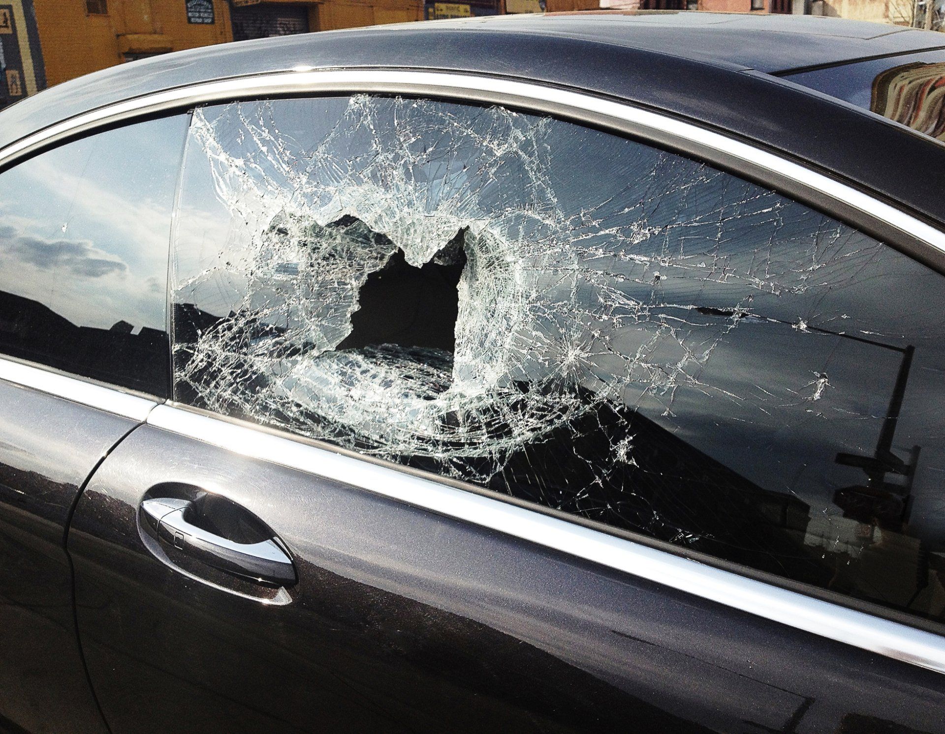 Damaged car window with a large hole, likely from vandalism. The vehicle is dark-colored, parked outdoors.