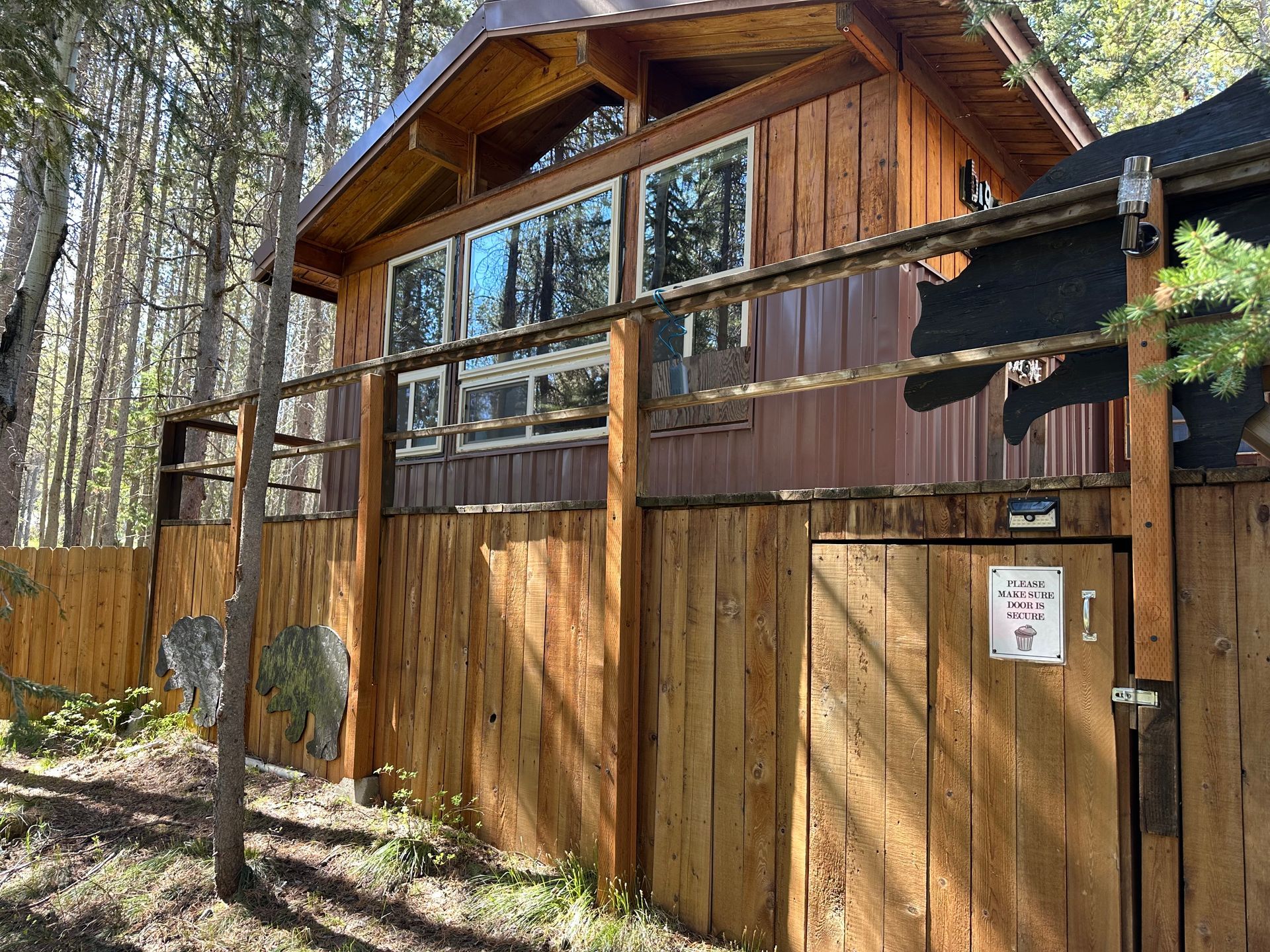 Wooden cabin with a fence in a forest, brown and tan hues, sunny day.