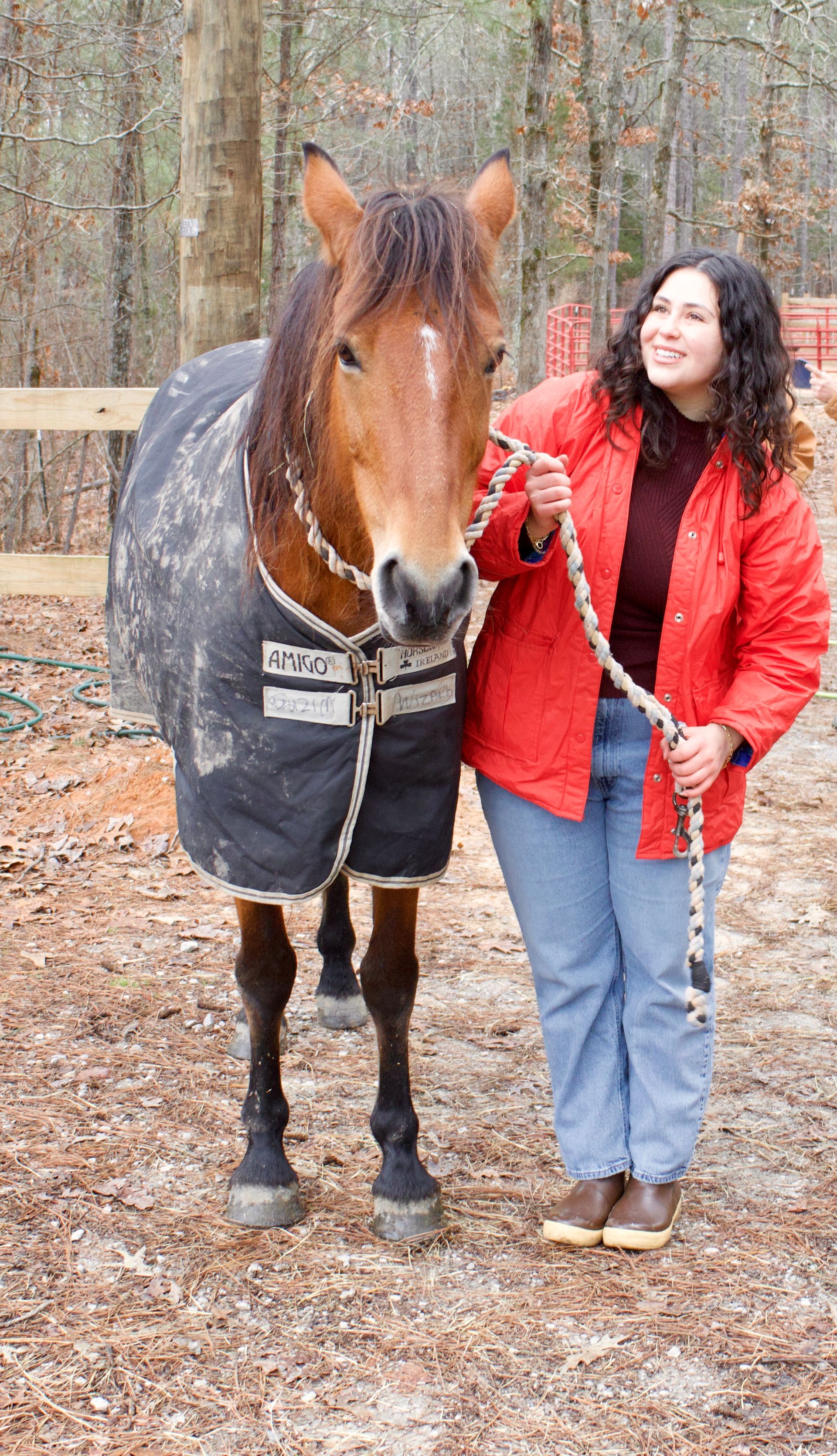 A woman in a red jacket is standing next to a brown horse.