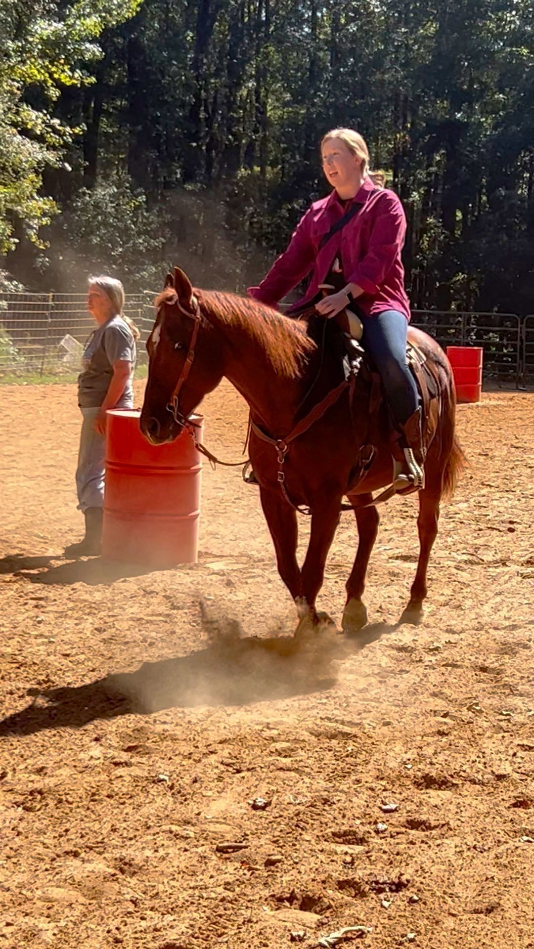 Woman on a brown horse riding around barrels in a dirt arena, another person watches.