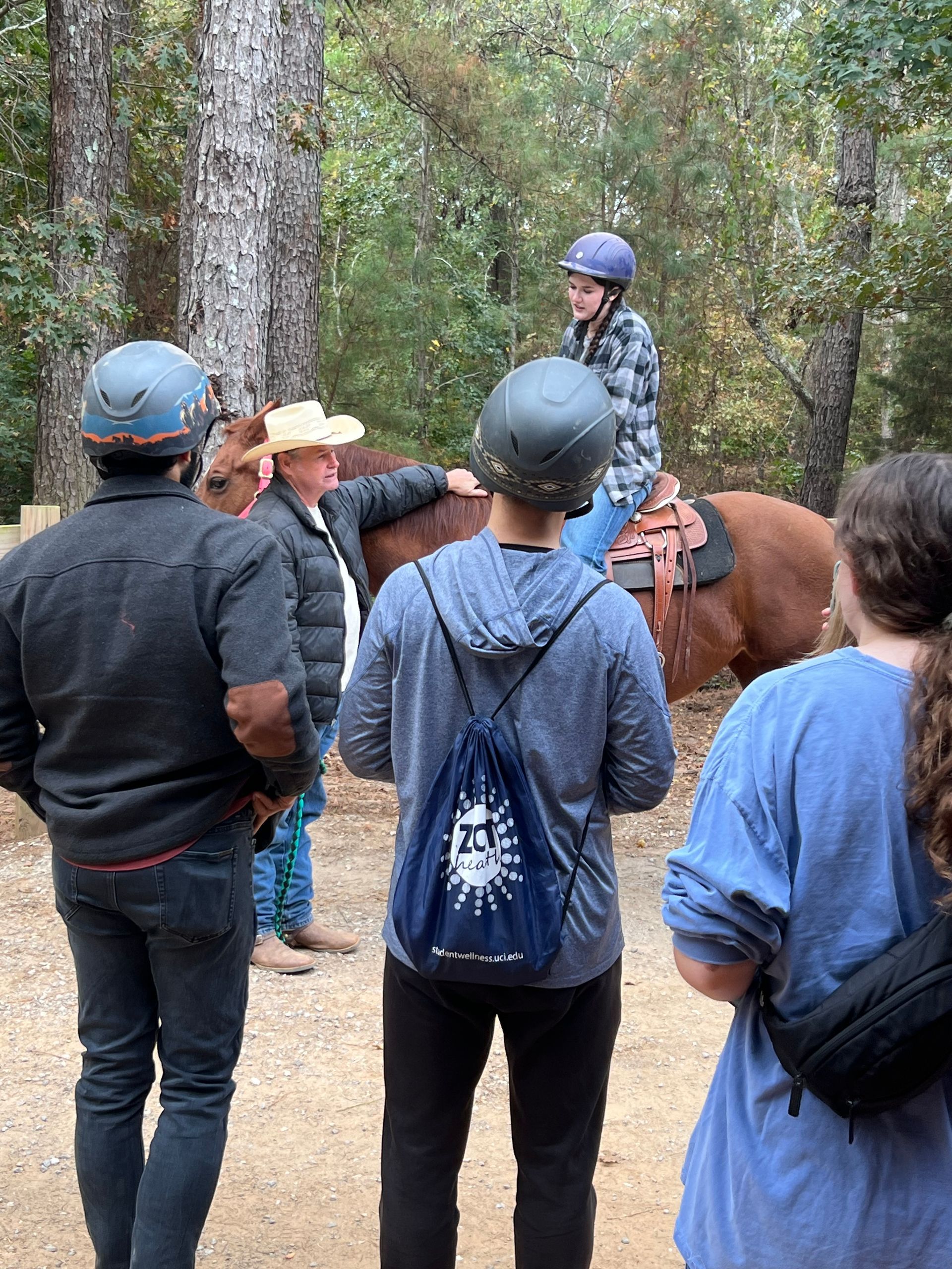 A person on a horse is watched by several people. Outdoors near trees; all wearing helmets.
