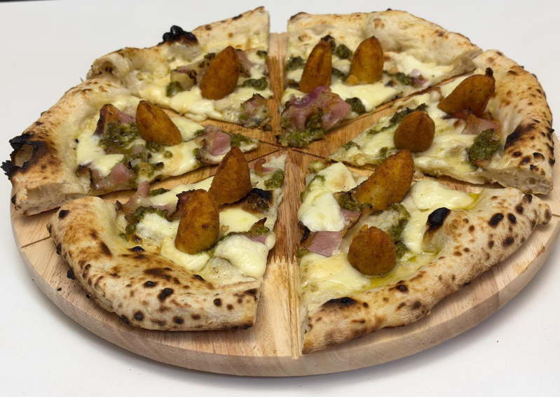 Pizza con formaggio bianco, prosciutto, pesto verde e crocchette di patate dorate su un tagliere di legno.