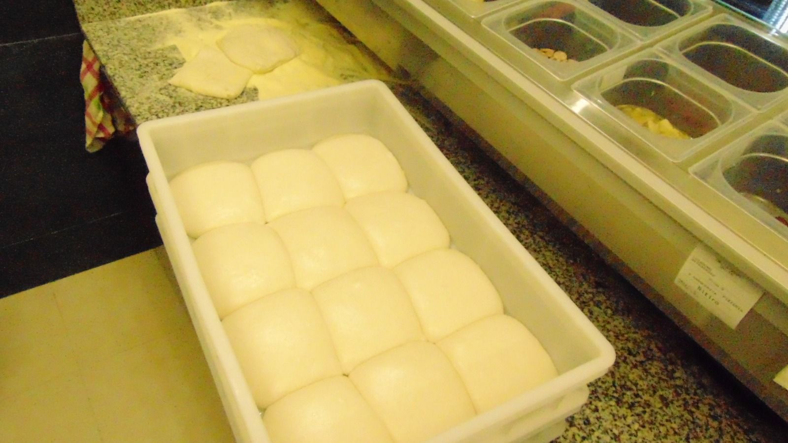 Contenitore bianco pieno di palline di impasto per pizza preparate, accanto a un piano di lavoro con condimenti.