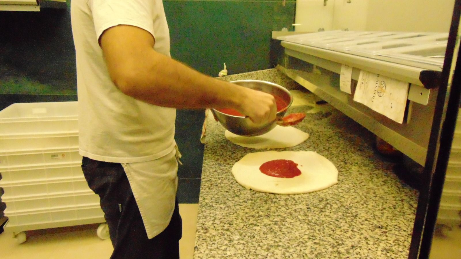 Una persona con un grembiule bianco spalma la salsa di pomodoro sull'impasto della pizza in cucina.