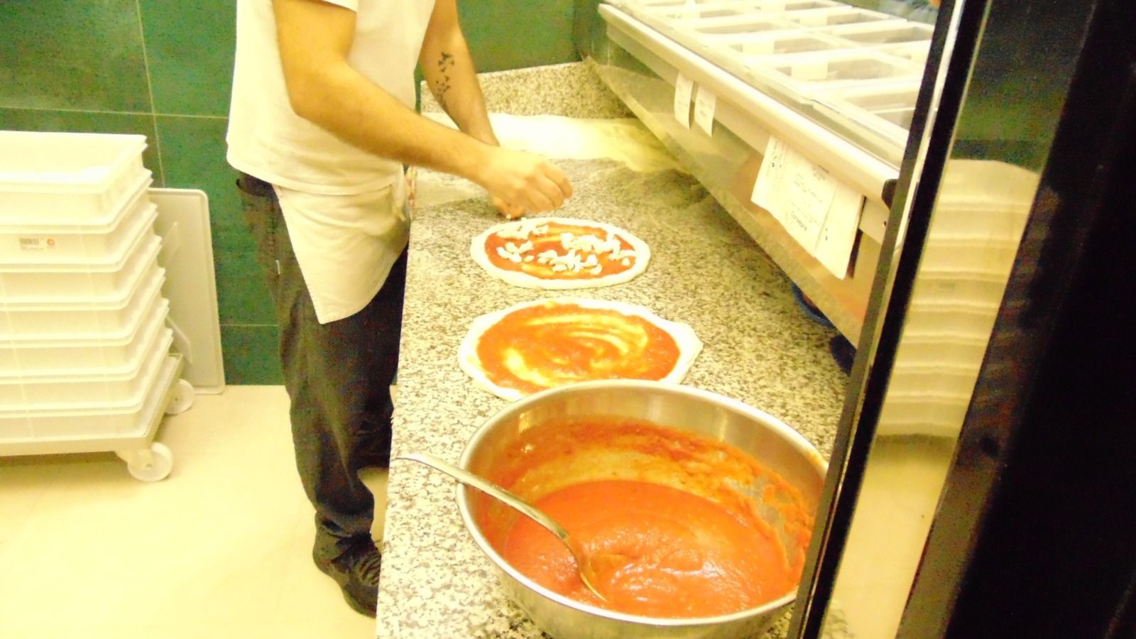 Persona che prepara la pizza in cucina, utilizzando salsa di pomodoro e altri condimenti.