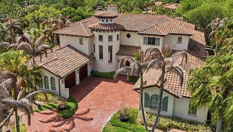 Aerial view of a large Mediterranean-style villa with red tile roofs, palm trees, and a brick driveway