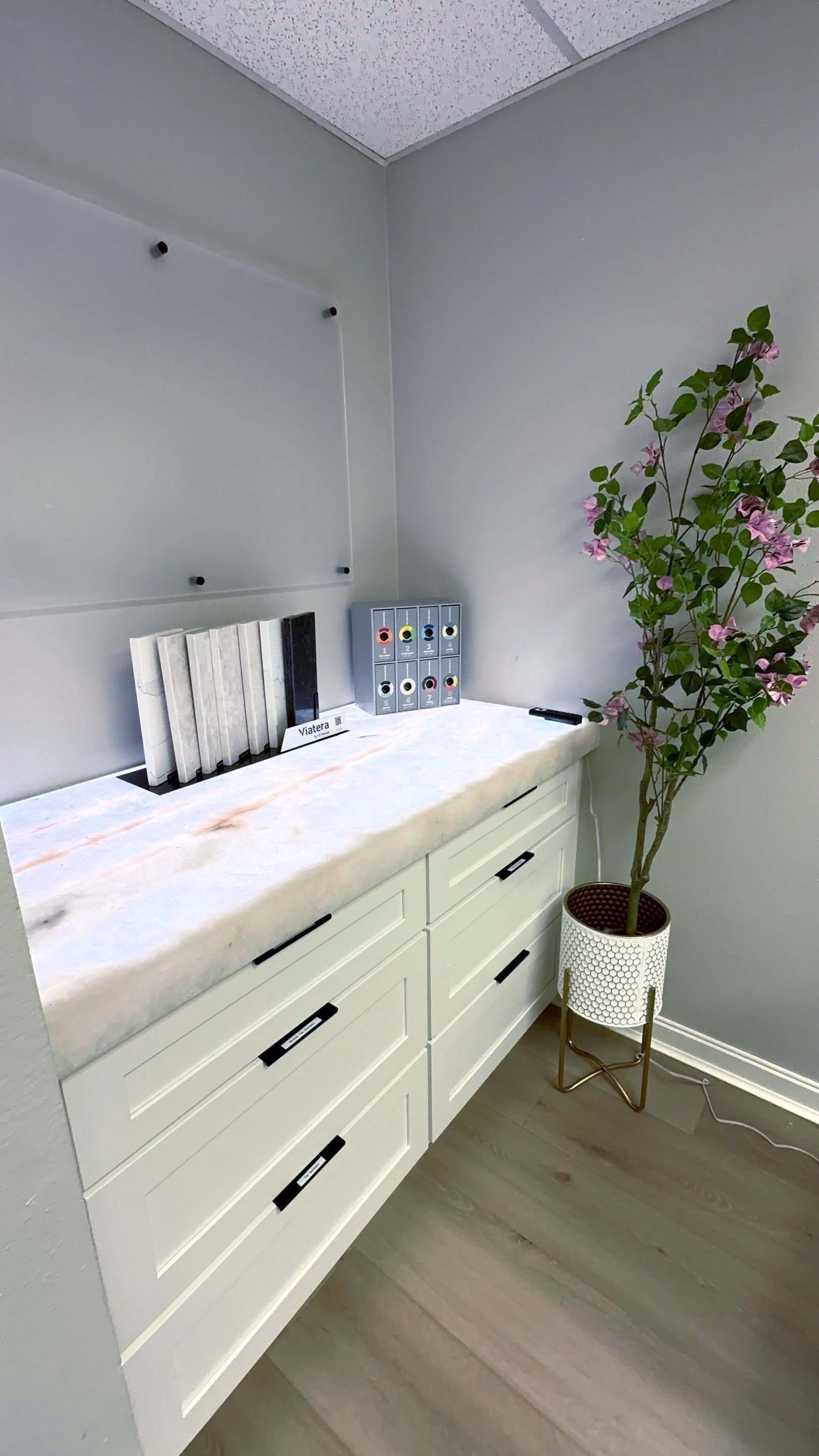 White changing table with drawers in a light gray nursery, beside a potted flowering plant and a wall shelf