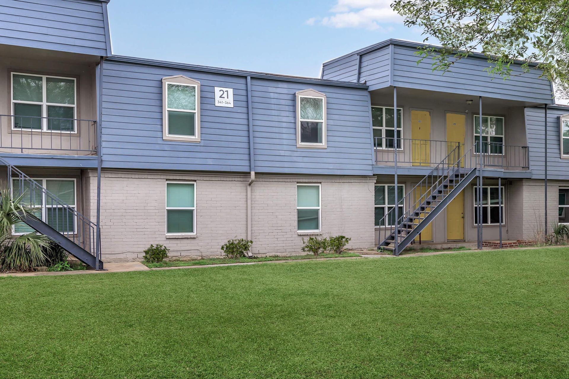 Two-story blue apartment building exterior with yellow doors, stairs, and a green lawn.