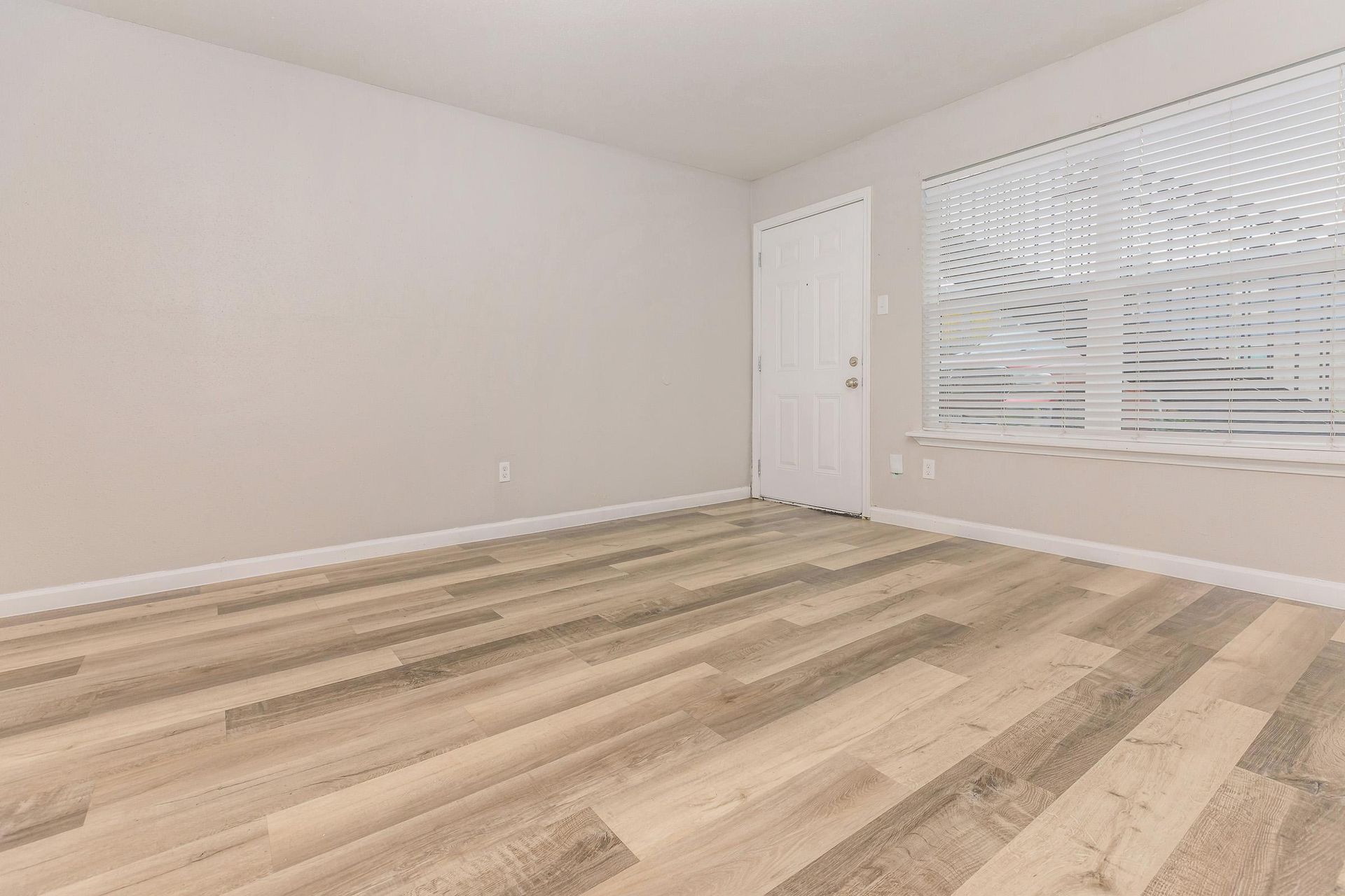 Empty apartment room with wood-look flooring, beige walls, a window with blinds, and a white door.