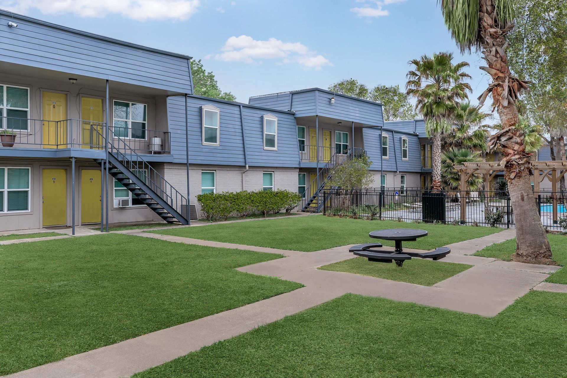 Exterior view of a blue apartment community with yellow doors, stairs, palm trees, and green lawn.