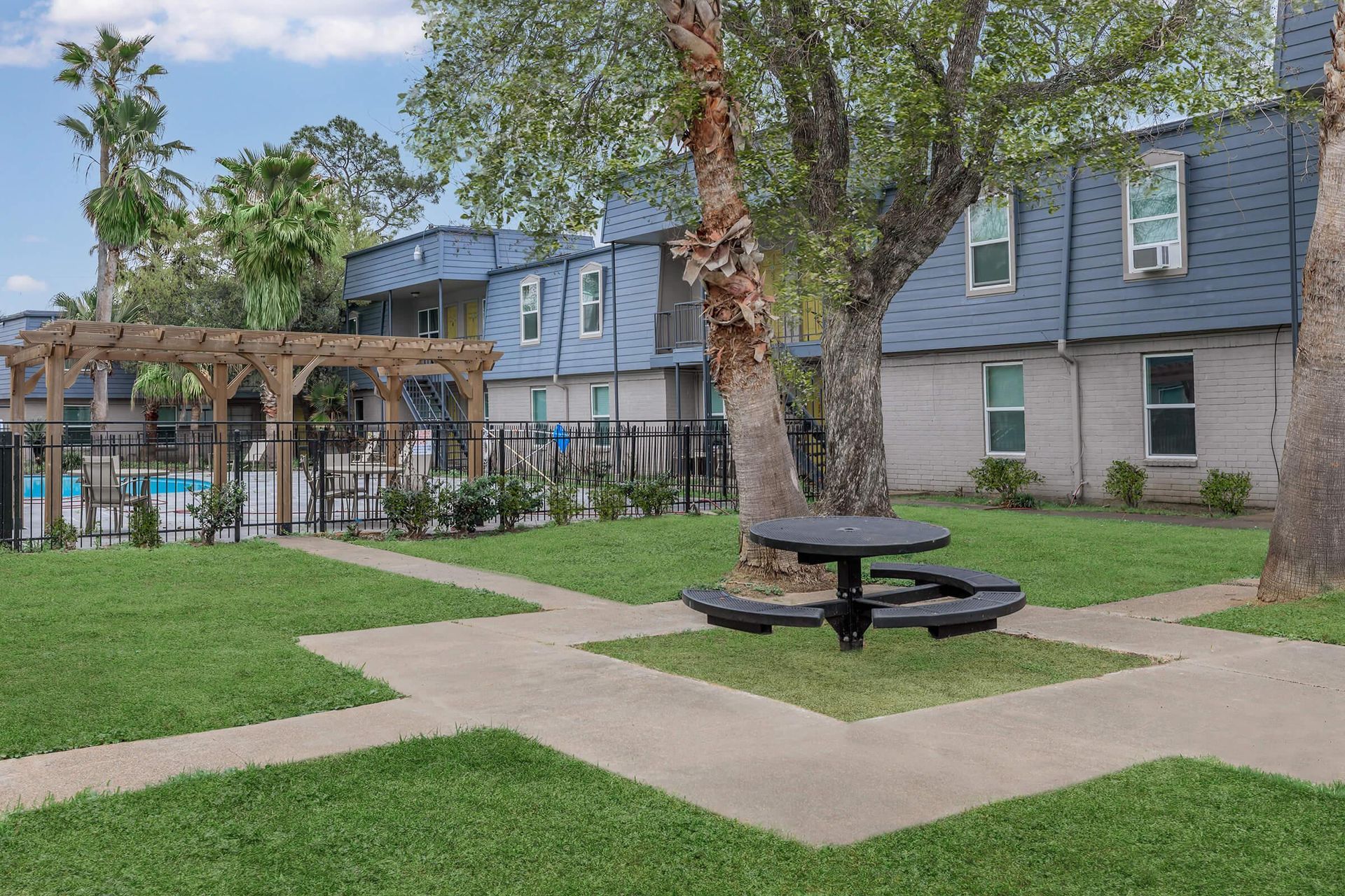 Outdoor community courtyard with palm trees, a circular picnic table, and a fenced pool area.