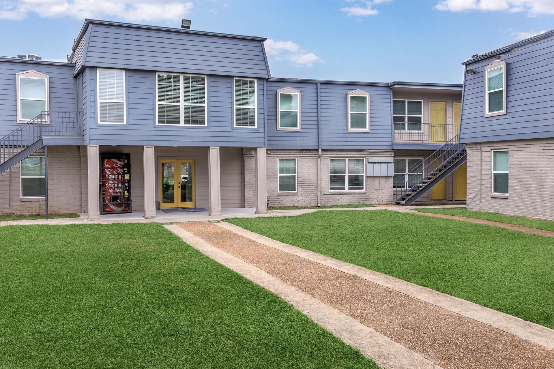 Exterior view of a blue, two-story apartment building with yellow doors and a grassy courtyard.