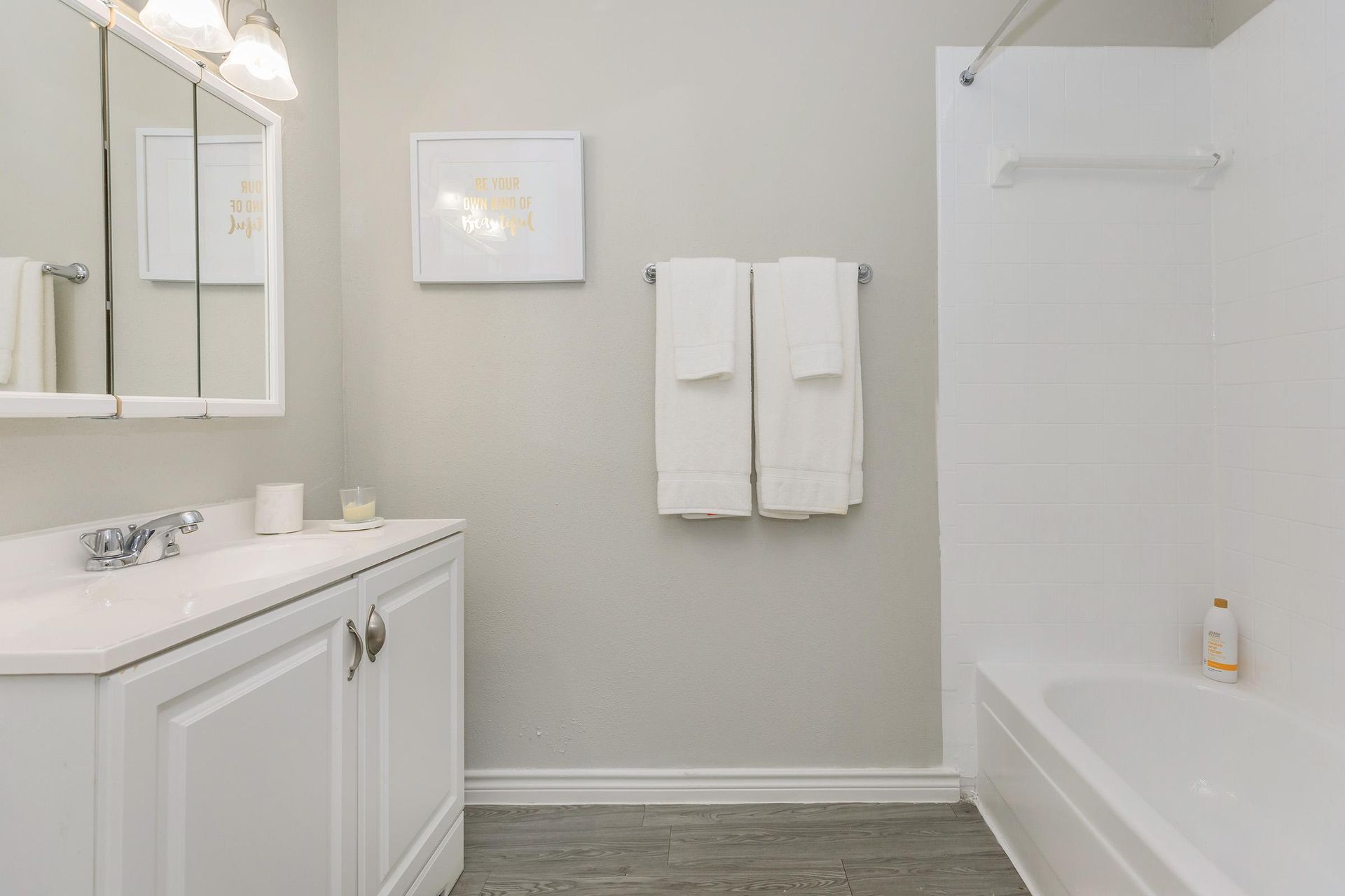 White apartment bathroom with vanity, mirror, towel rack, and bathtub/shower.