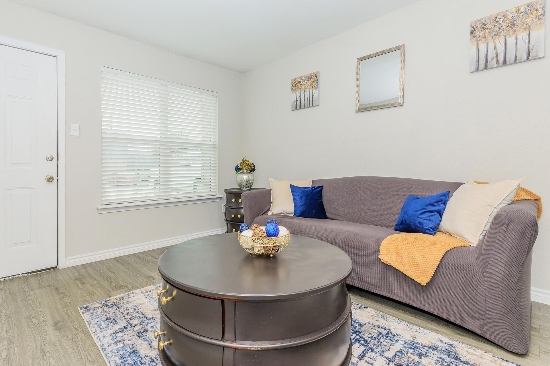 Living room in an apartment featuring a gray sofa, round dark coffee table, and window blinds.
