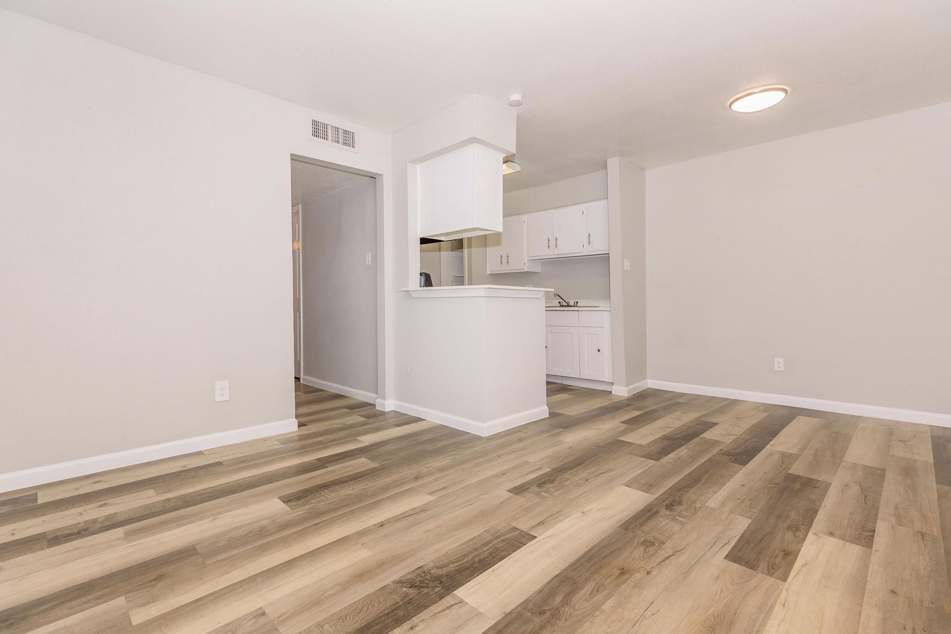 Living area with kitchen in an unfurnished apartment, white cabinets and light wood flooring.