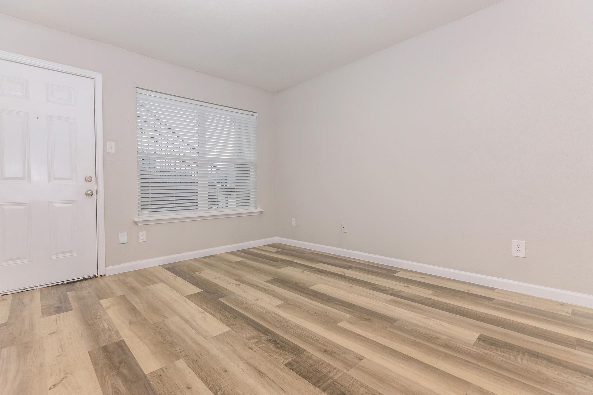 Empty apartment room with light wood vinyl flooring, neutral walls, and a window with blinds.