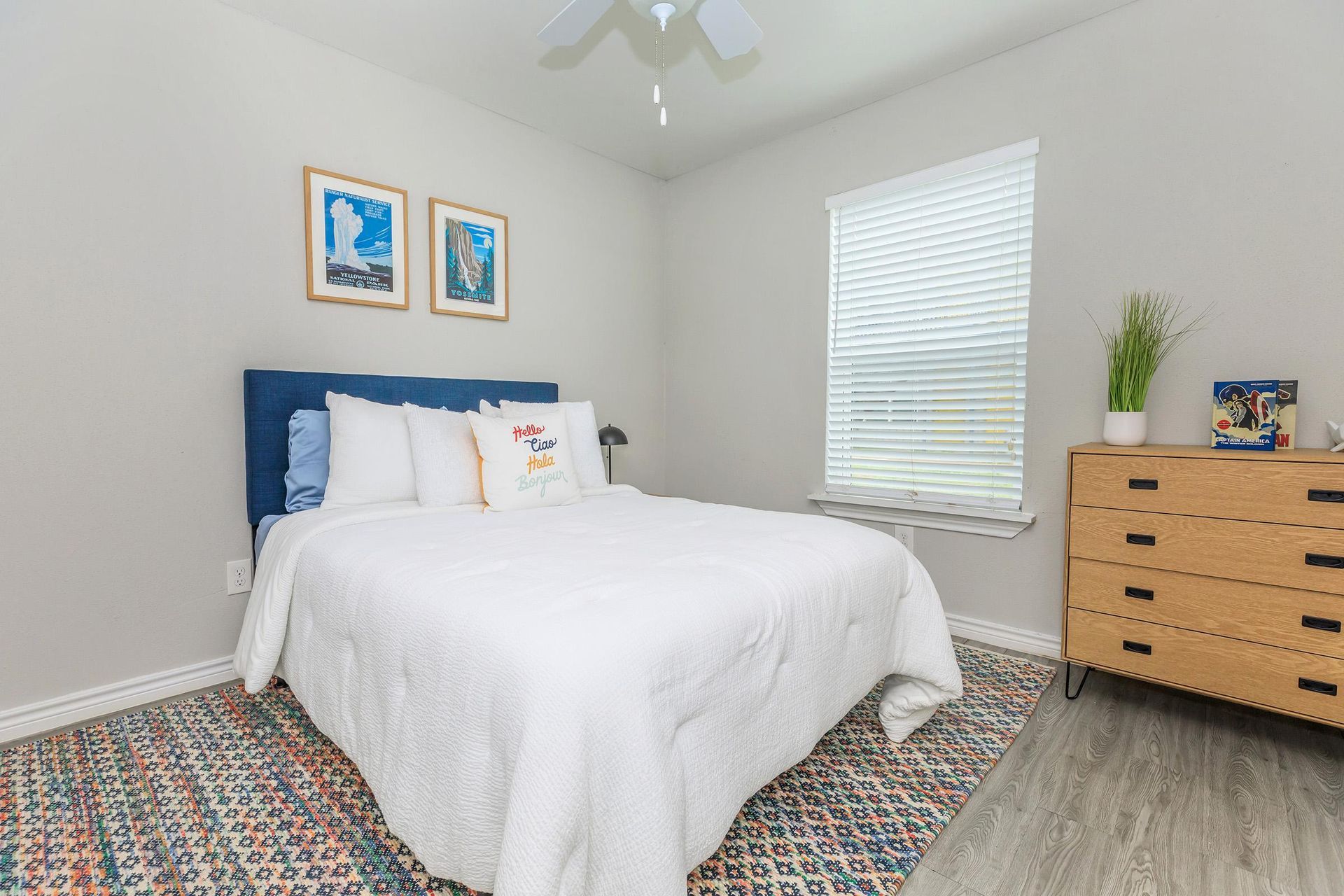 Bedroom in apartment with white bedding, blue headboard, dresser, and window blinds.