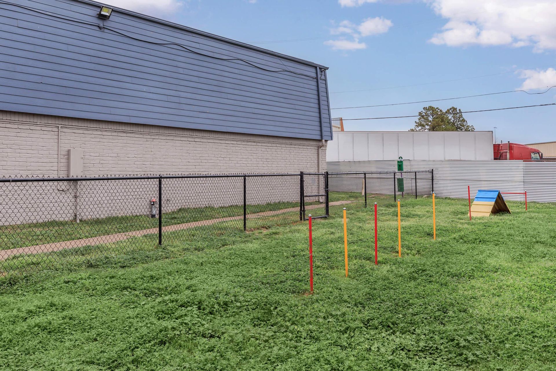 Fenced grassy community area with colorful posts and a small blue ramp near a beige building.