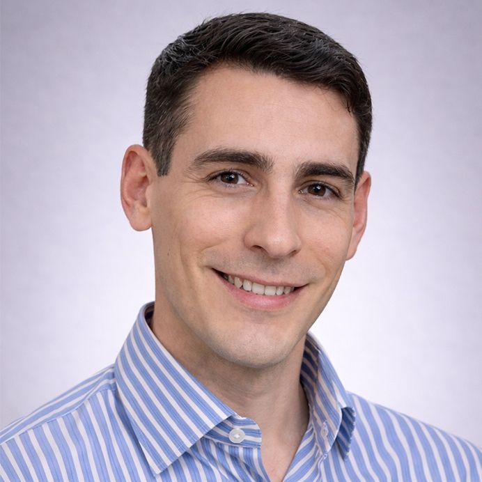 A headshot of a person wearing a dark blazer and a light blue-and-white striped shirt, set against a circular orange background.