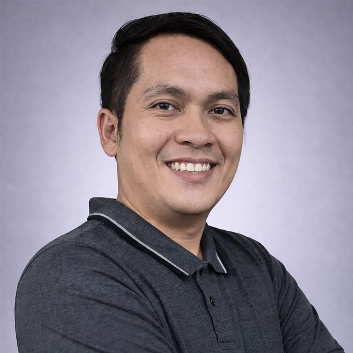 A person with short dark hair and glasses smiles, wearing a dusty rose shirt, set against a circular orange background.