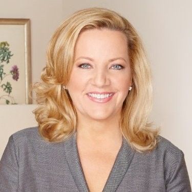 A person with short, gray hair, wearing a black shirt and a pendant necklace, smiling against a light gray background.