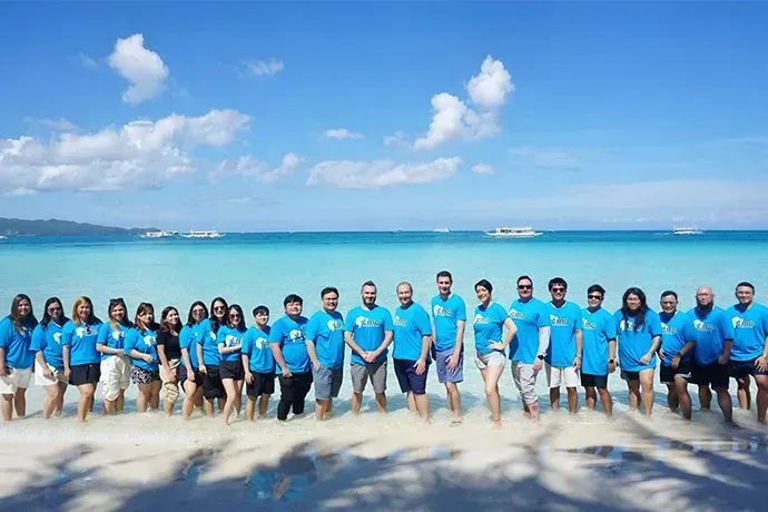 A large group of people in matching blue shirts stands in a line on a white sand beach by turquoise water under a blue sky.
