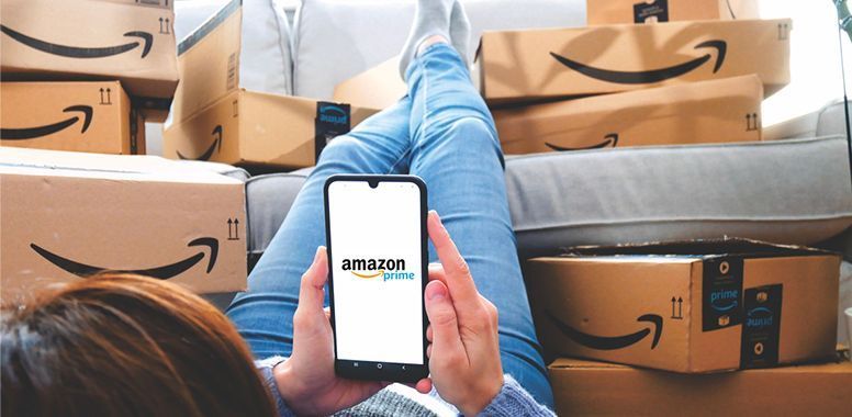 A person holds a phone displaying the Amazon Prime logo, surrounded by several Amazon shipping boxes.