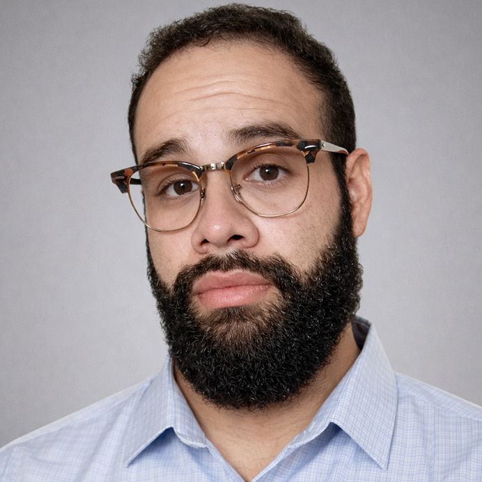 A person with a full beard and glasses, centered in a circular frame against a solid mustard-yellow background.