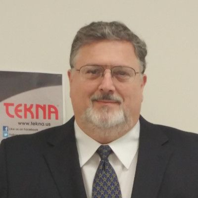 A person with short, gray hair, wearing a black shirt and a pendant necklace, smiling against a light gray background.