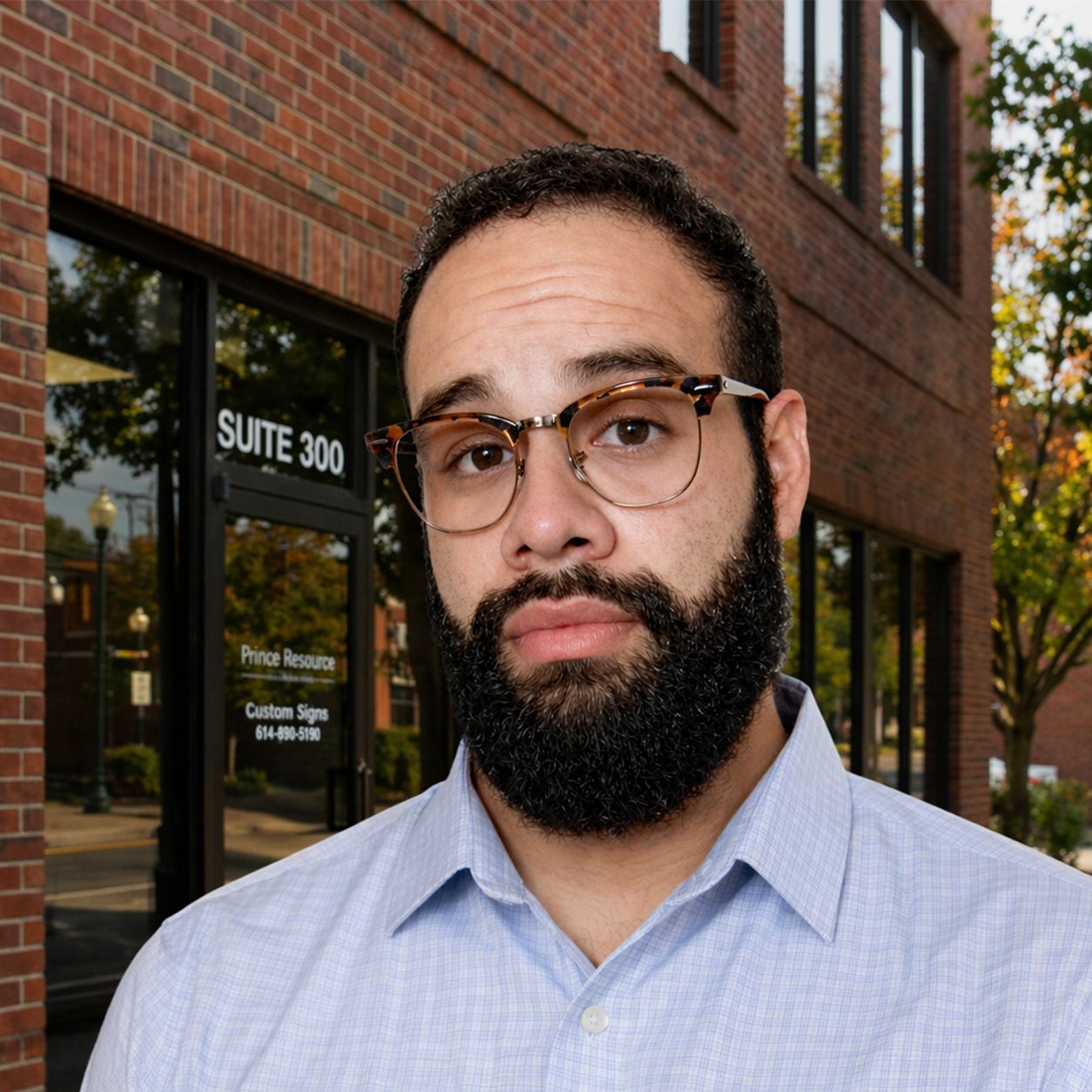 Portrait of a bearded person wearing glasses and a light blue shirt against a gray background