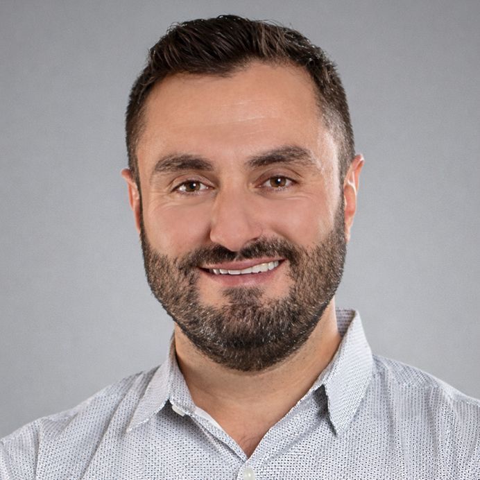 A person with short, gray hair, wearing a black shirt and a pendant necklace, smiling against a light gray background.