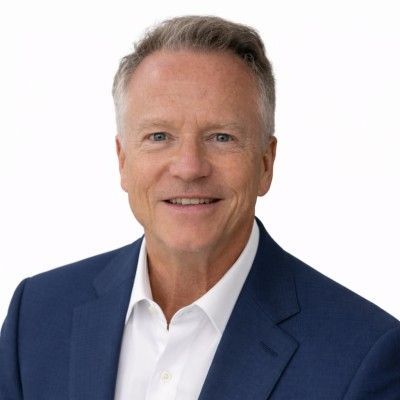 A person with short, gray hair, wearing a black shirt and a pendant necklace, smiling against a light gray background.