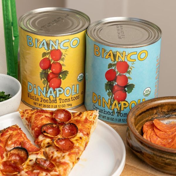 Two cans of Bianco DiNapoli tomatoes, one yellow and one blue, near a plate of pepperoni pizza and a bowl of pepperoni.