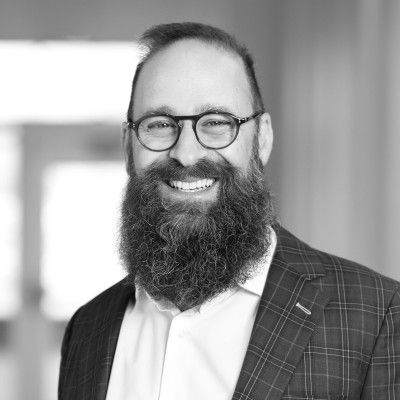 A person with short, gray hair, wearing a black shirt and a pendant necklace, smiling against a light gray background.