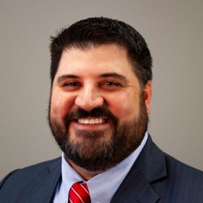 A person with short, gray hair, wearing a black shirt and a pendant necklace, smiling against a light gray background.