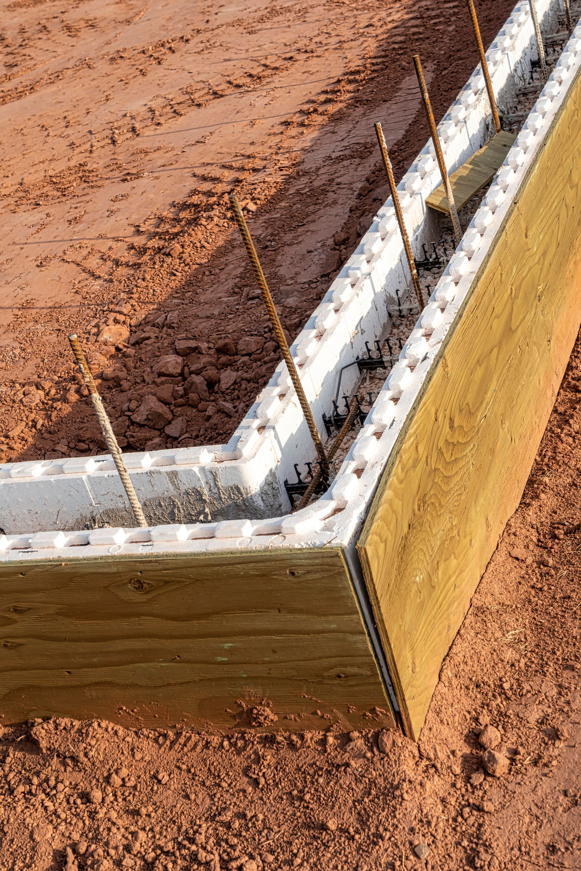 Corner of an ICF foundation form with rebar and brown insulation, set on red earth.