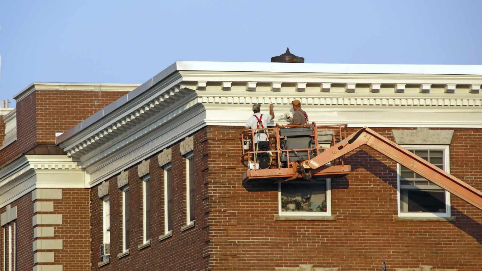 two men are painting the roof of a brick building in sunny weather. 