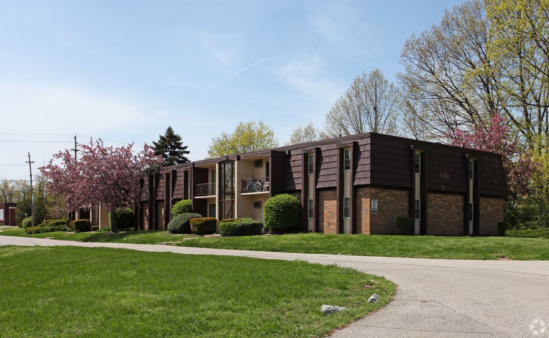 Apartment building with brown roof and brick exterior. Green lawn, walkway, and trees in spring.