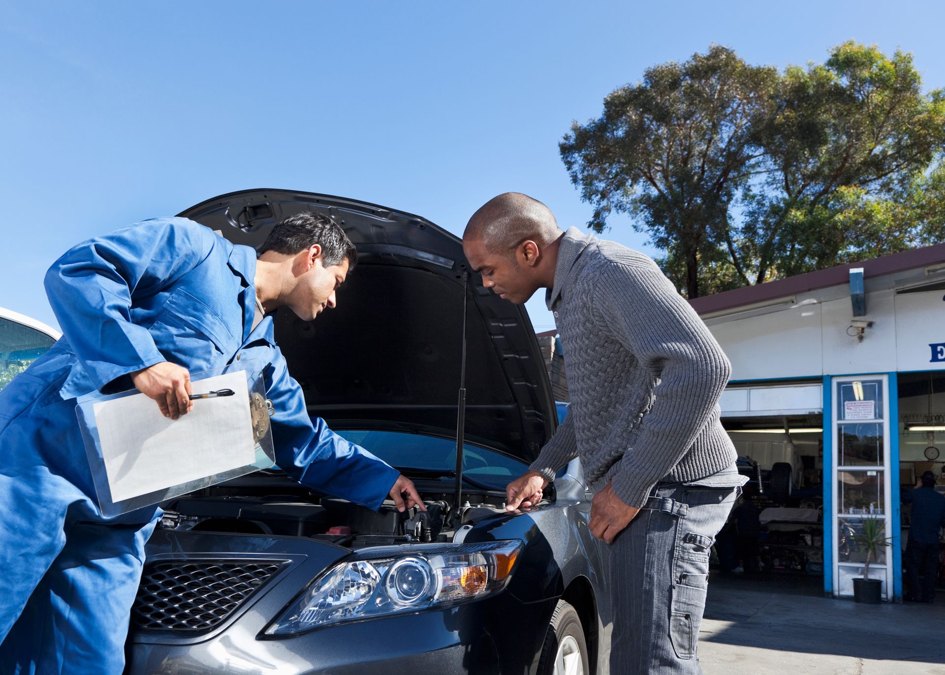 Mechanic in blue overalls and customer inspecting car engine outside a garage.