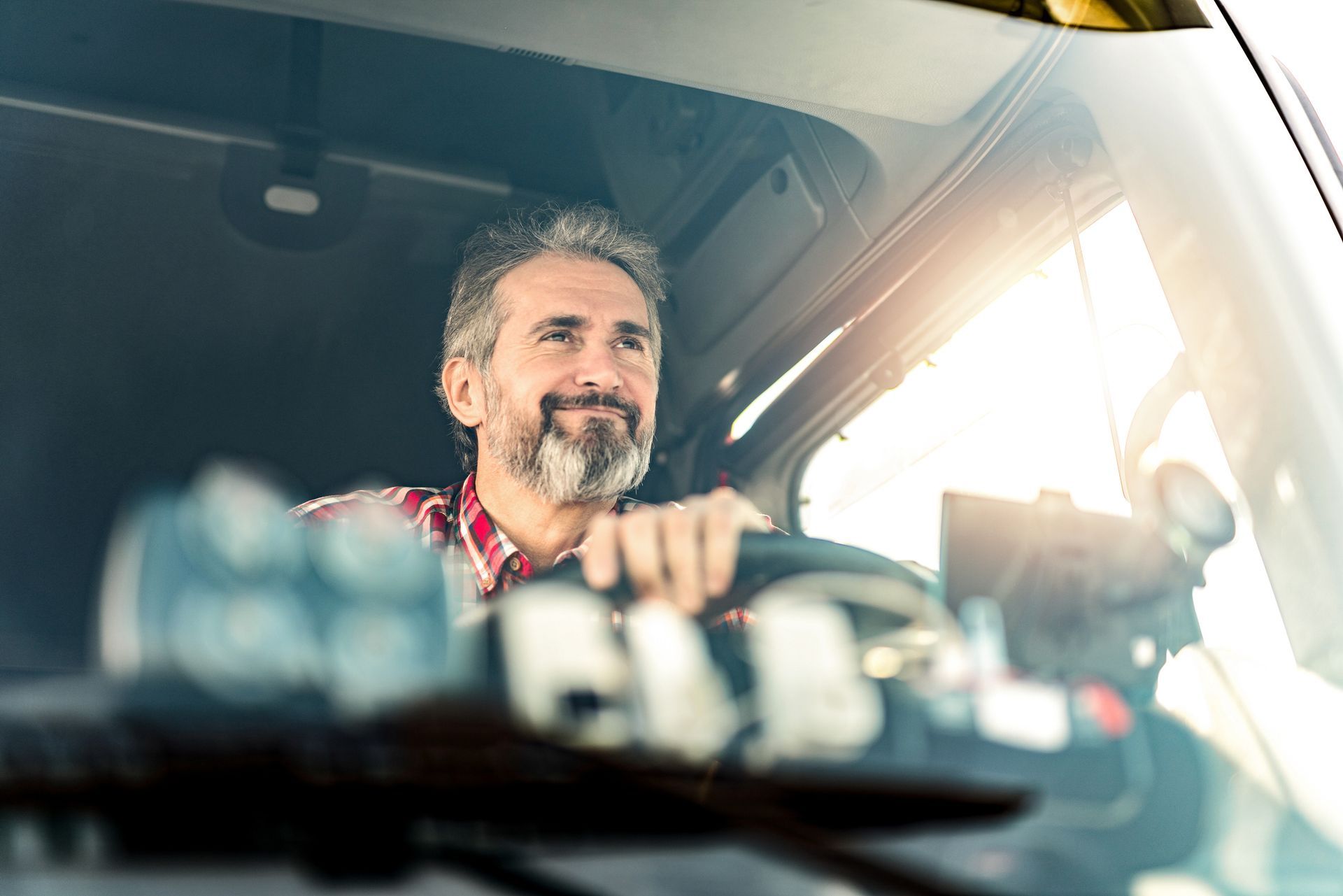 Man with gray beard smiles while driving a semi-truck, seen from inside the cab.