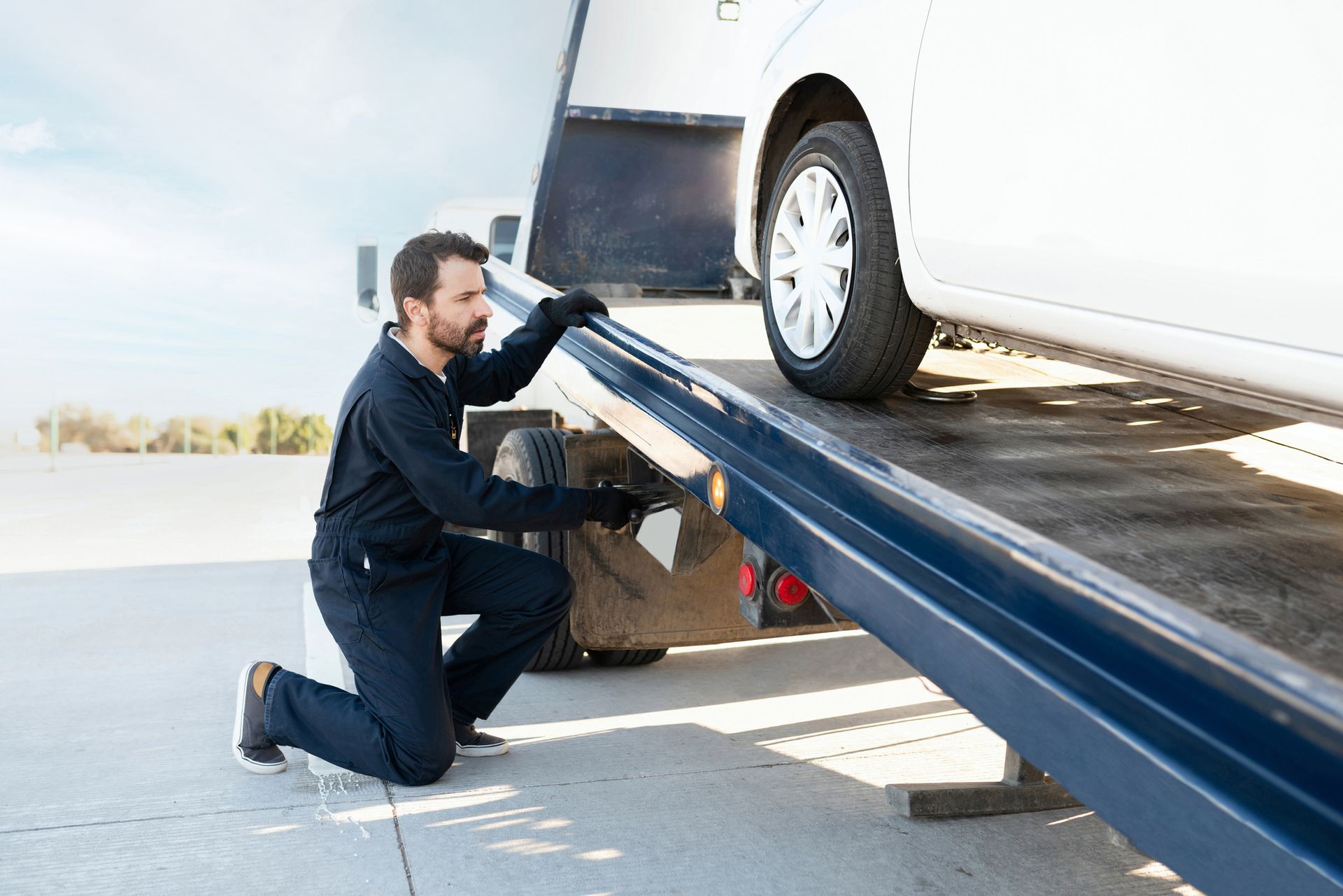 A tow truck operator secures a white car onto a flatbed ramp outdoors.