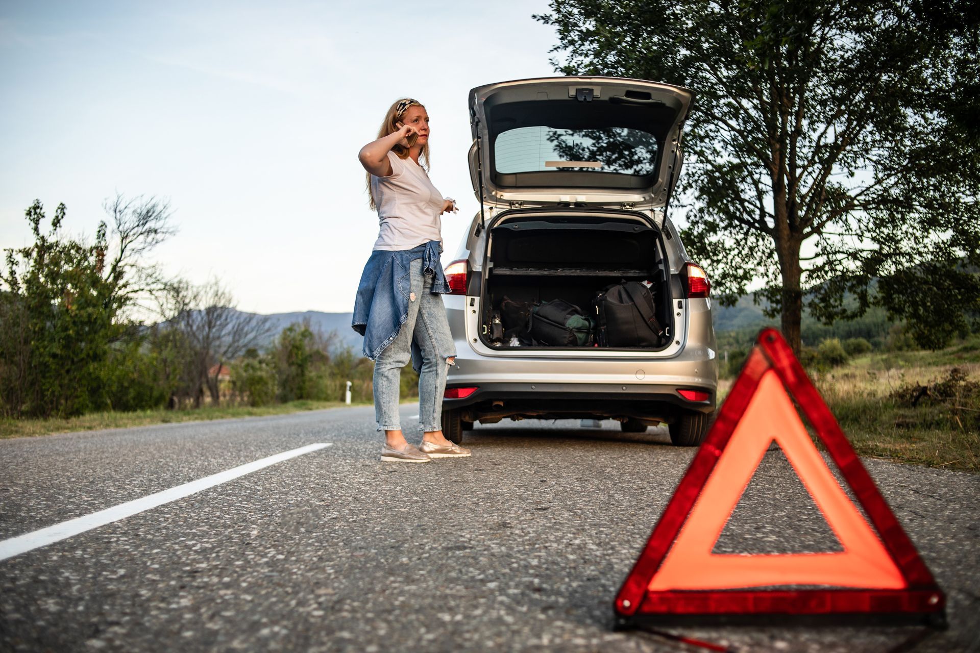 Woman on phone by a broken-down car with trunk open, next to a roadside warning triangle.