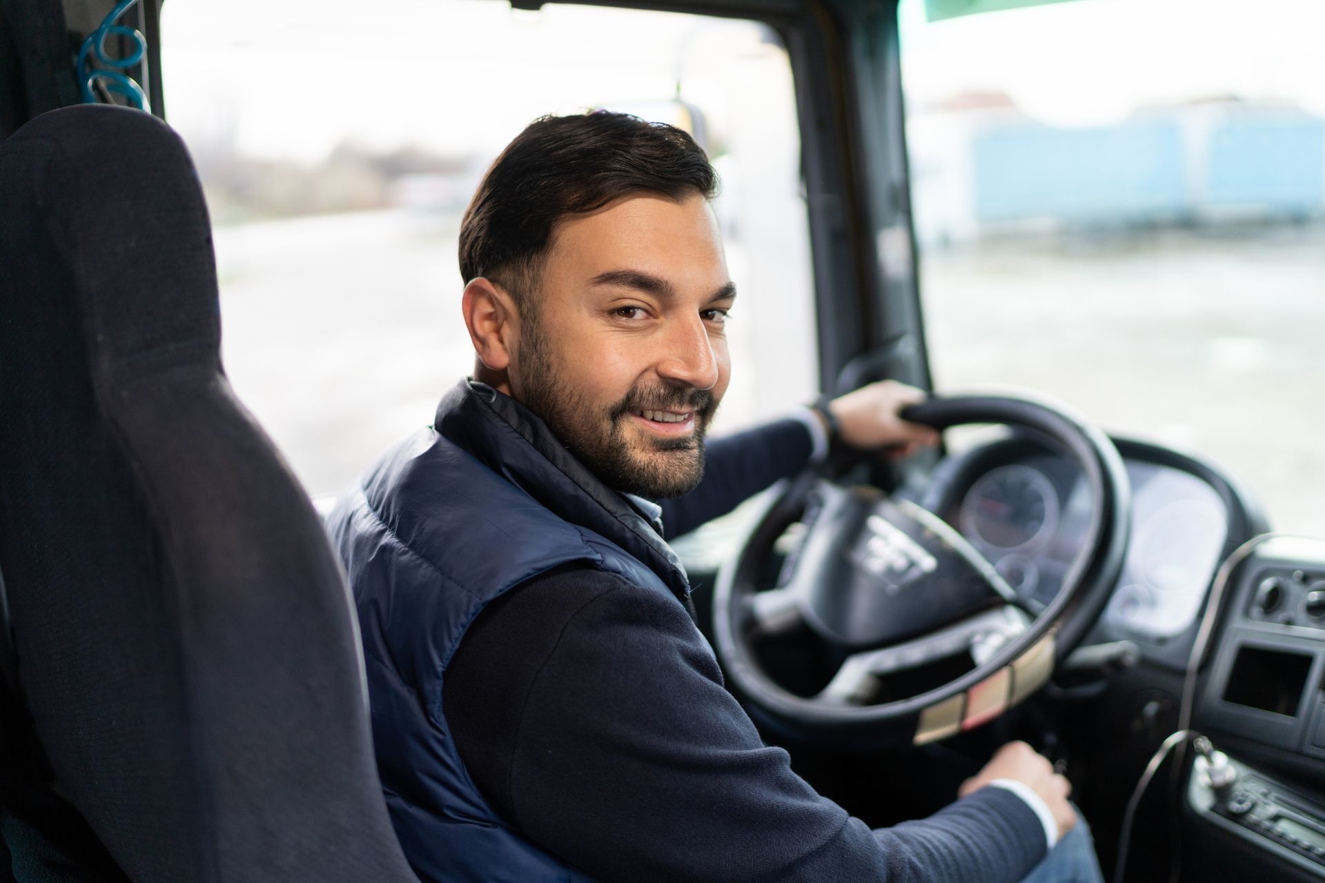 Man smiling, driving a truck. He’s wearing a vest, looking over his shoulder in a vehicle.