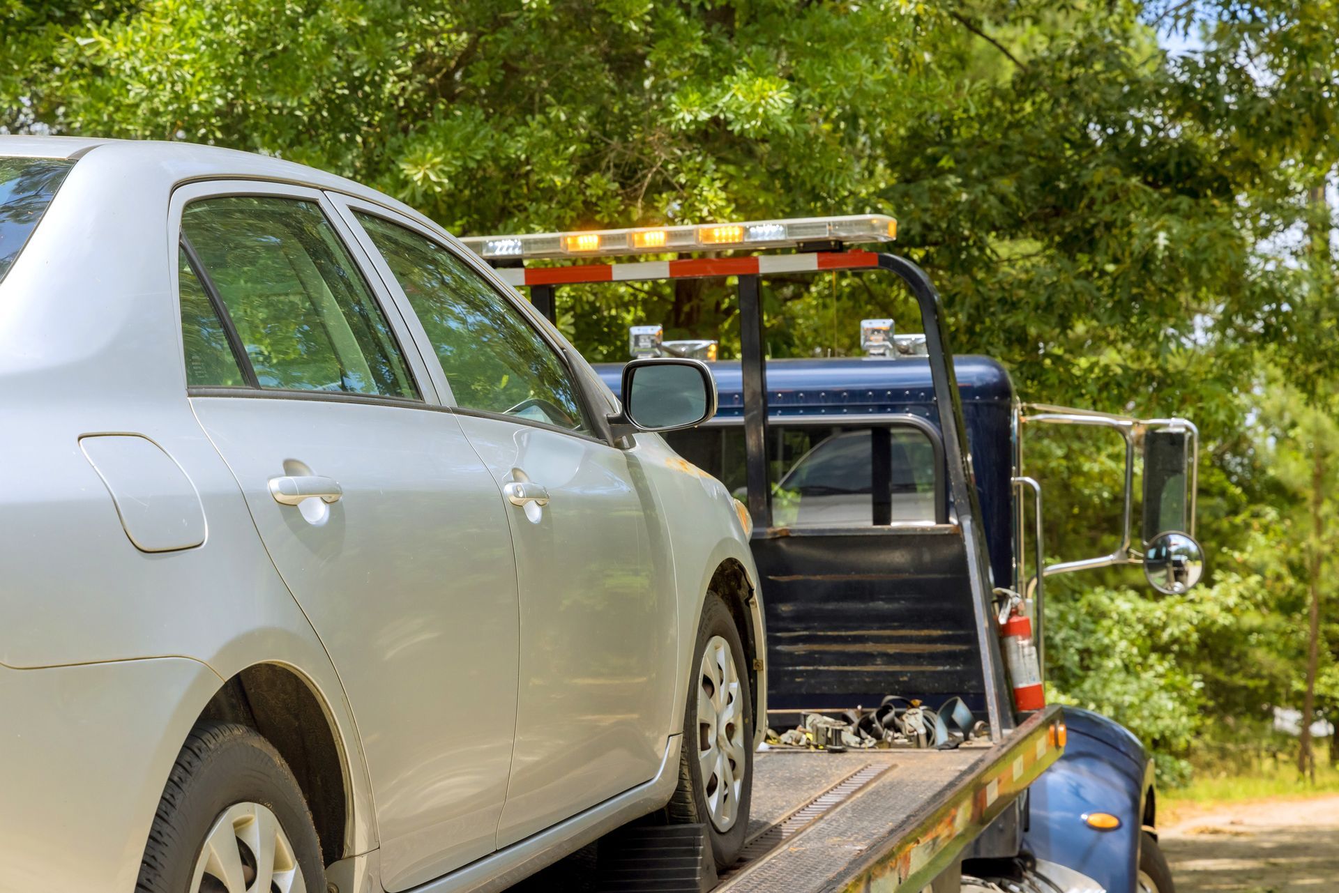 Silver car being towed on a flatbed tow truck; green trees in background.