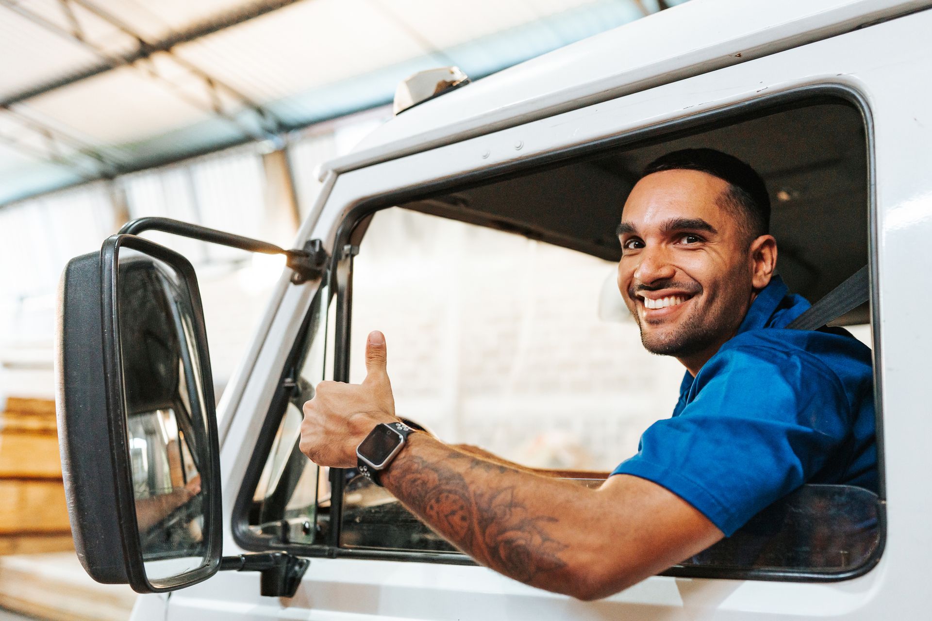 Man in a truck cab gives a thumbs-up, smiling. Wearing blue uniform, watch, bright setting.