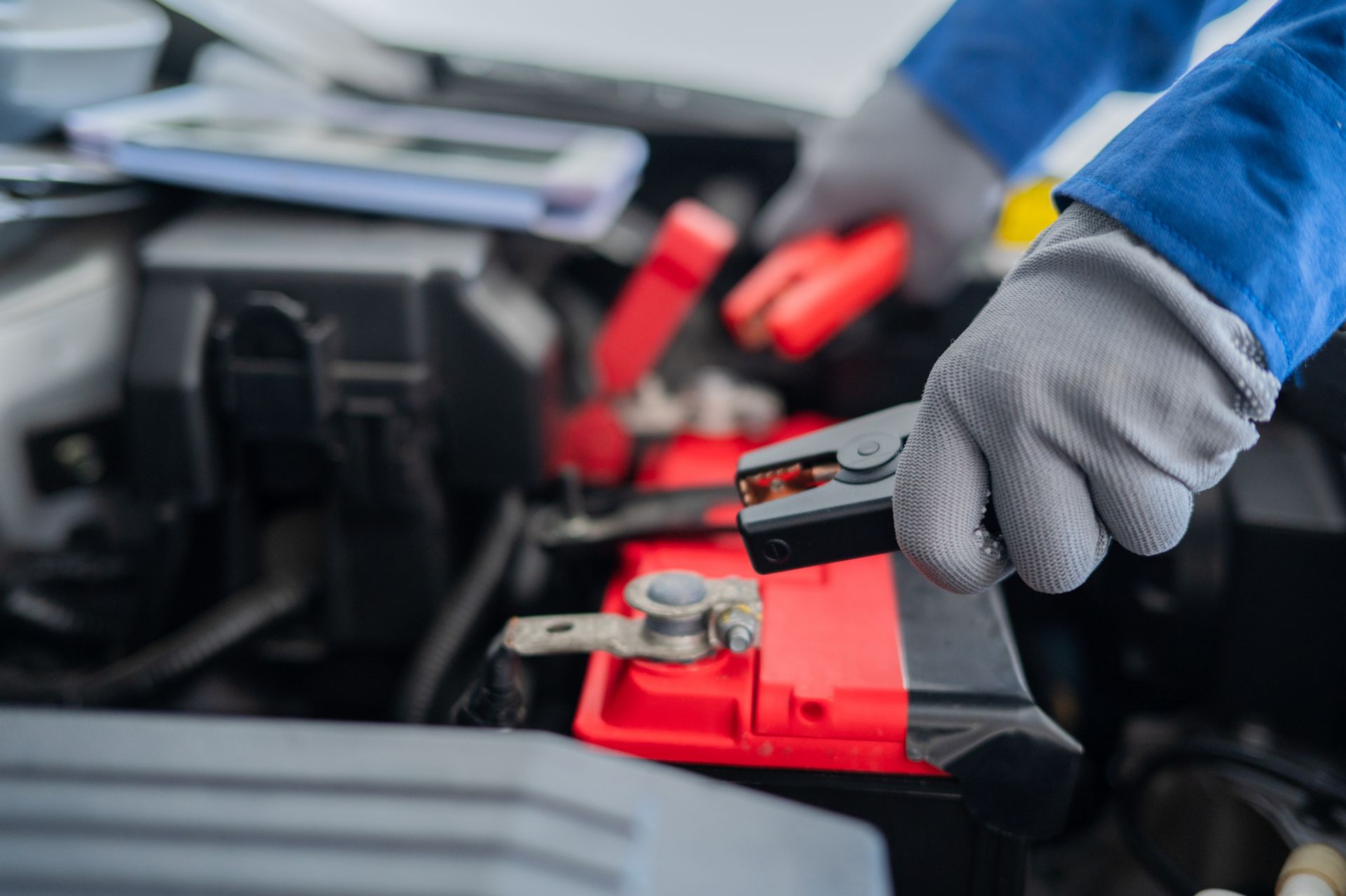 Mechanic's hands wearing gloves attaching jumper cables to a red car battery in an engine bay.